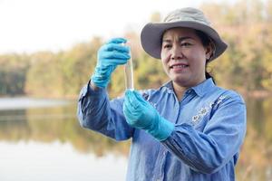 Asian women environment researcher holds tube of sample water to inspect at the lake. Concept, explore, analysis water quality from natural source. Ecology field research. photo