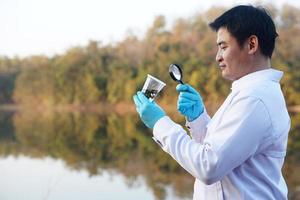 Asian man ecologist holds magnifying glass to inspect water in transparent glass from the lake. Concept, explore, analysis water quality and creature from natural source. Ecology field research. photo