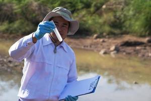 Asian man environment researcher holds tube of sample water to inspect at the lake. Concept, explore, analysis water quality from natural source. Ecology field research. photo