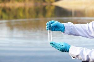 las manos ecologistas de primer plano usan guantes azules y sostienen un tubo de vidrio de prueba que contiene agua de muestra en el lago. concepto, explorar, inspeccionar la calidad del agua de fuente natural.