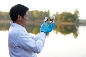 Asian man ecologist holds magnifying glass to inspect water in transparent glass from the lake. Concept, explore, analysis water quality and creature from natural source. Ecology field research. photo