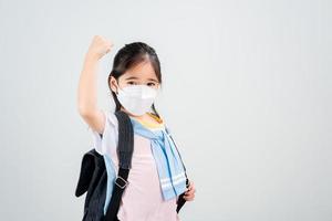 niña asiática lleva una mochila con máscara de respirador para proteger el brote de coronavirus y apunta con la mano al fondo en blanco, nuevo virus covid-19 foto
