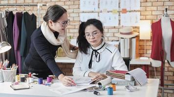 Asian middle-aged female fashion designer teaches a young teen trainee tailor in studio with colorful thread and sewing fabric for dress design collection ideas, professional boutique small business. photo
