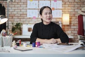 Asian mature female fashion designer is sitting at table, looking at camera and in studio, working with creative and sewing for dress design collection, professional boutique tailor SME entrepreneur. photo