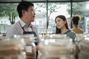 Asian male shopkeeper describes natural organic products to White woman customer in refill store, zero-waste and plastic-free grocery, environment-friendly, sustainable lifestyles in reusable shop. photo