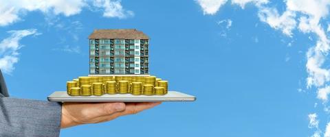 Businessman holding a tablet with piles of golden coins and condominium on white cloud and blue sky background photo