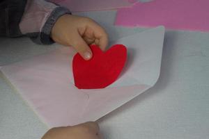 niño sosteniendo una carta con un corazón de papel rojo, una carta de amor para el día de San Valentín, un regalo para el día de la madre, una niña puso un corazón hecho a mano dentro de un sobre, saludos navideños de su hija, maqueta para deseos de amor. foto