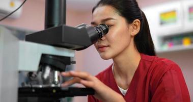 Close up shot, Asian young veterinarian female wearing uniform sitting and looking through a microscope, her hand slide sample of animals while working in lab, video