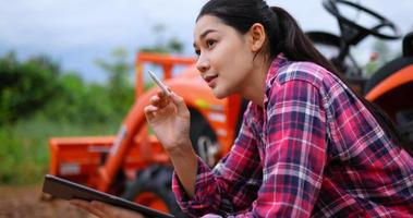 mujer agricultora muy joven sentada para comprobar la calidad de los productos agrícolas en el tractor, mirando a su alrededor y usando un lápiz óptico digital para escribir en la tableta, trabajando en la plantación cuando hace calor video