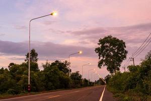 una vista de la luz de una farola naranja en una carretera mojada después de la lluvia con árboles en el campo tailandés. foto