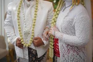 hermosos novios de pie mientras usan un vestido de novia con un collar de flores de jazmín y magnolia para una ceremonia de boda tradicional en indonesia foto