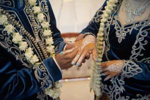 el novio pone el anillo de bodas en la mano de la novia con un vestido de novia con un collar de flores de jazmín y magnolia en una ceremonia de boda tradicional en indonesia foto