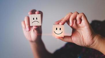 Smile face on the bright side and sad face on the dark side on wooden block cube for positive mindset selection. Hand showing mental health and emotional state concept.