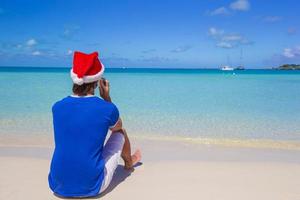 vista trasera del joven con sombrero de santa con teléfono en la playa caribeña tropical foto