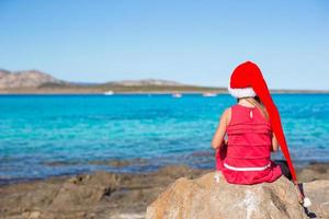 adorable niña con sombrero de santa y vestido rojo sentada en una gran piedra foto