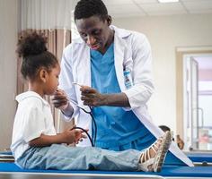 Selective focus at doctor person playing with cute afro child patient in hospital ward. Friendly pediatrician entertaining girl kid enjoying with stethoscope for heartbeat during medical exam. photo