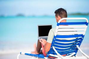 Young man with laptop on tropical caribbean beach. Man sitting on the sunbed with computer and working on the beach photo