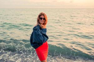 Outdoor fashion portrait of stylish girl wearing trendy sunglasses and jeans jacket on the beach. Hands up, retro filter, flecks of sunlight photo