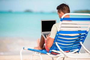 Young man with laptop on tropical caribbean beach. Man sitting on the sunbed with computer and working on the beach photo