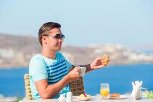 chico joven desayunando en un café al aire libre con una vista increíble de la ciudad de mykonos. hombre tomando café en la terraza del hotel de lujo foto