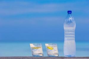 Glasses of waters with lemon and bottle on wooden on sea background photo