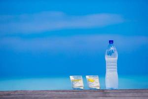 Glasses of waters with lemon and bottle on wooden on sea background photo