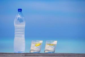 Glasses of waters with lemon and bottle on wooden on sea background photo