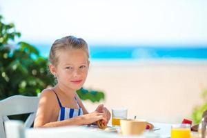 Adorable little girl having breakfast at cafe with sea view photo