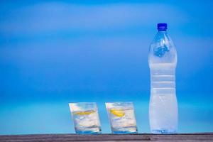 Glasses of waters with lemon and bottle on wooden on sea background photo