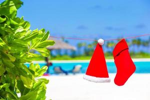 sombrero rojo de santa y calcetín de navidad colgando en la playa tropical foto