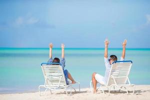Two happy people having fun on the beach, sitting on comfortable sunbed with raised up hands for joy photo
