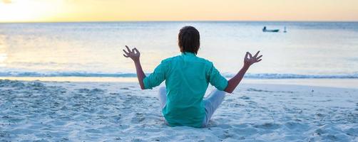 joven sentado en posición de loto en la playa de arena blanca foto