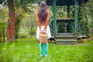 vista trasera de una joven en el jardín de botas de goma brillantes en manos de una cesta de paja con manzanas foto