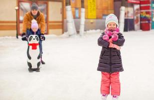 adorable niña en la pista de patinaje con padre y linda hermana en el fondo foto