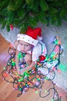 Little beautiful girl in Santa Claus hat sitting under the Christmas tree among garlands photo