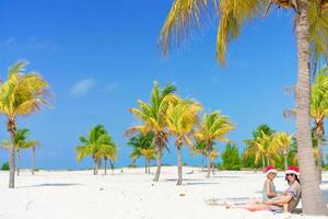 Young mom and little girl have fun at tropical beach in santa hat sitting near palm tree photo