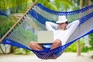 Young man with laptop at hammock on white beach photo