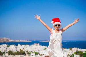 Little funny girl in red Santa hat outdoors background of Mykonos. Kid at street of typical greek traditional village with white walls and colorful doors on Christmas vacation in Greece photo