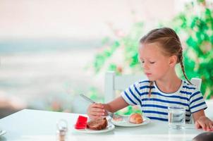 Adorable little girl having breakfast at cafe with sea view photo