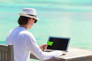 Young man working on laptop with credit card at tropical beach photo