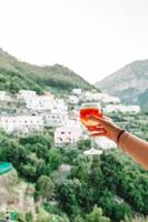 mano femenina sosteniendo un vaso con bebida de alcohol spritz aperol antecedentes de un hermoso y antiguo pueblo italiano en la costa de amalfi foto