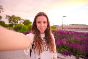 joven mujer caucásica haciendo selfie en el fondo de las atracciones al aire libre en la luz del atardecer foto