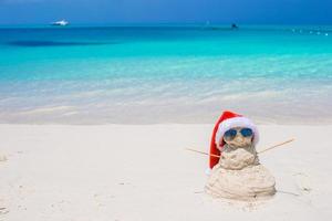pequeño muñeco de nieve de arena con sombrero rojo de santa en la playa caribeña blanca foto