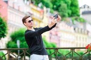 Tourist man taking travel photos with smartphone on summer holidays. Young attractive tourist taking selfie photo with mobile phone outdoors enjoying holidays travel destination in tourism.