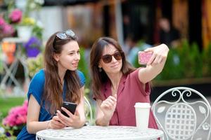 Two young girls taking selfie with smart phone at the outdoors cafe. Two women after shopping with bags sitting in openair cafe with coffee and using smartphone photo