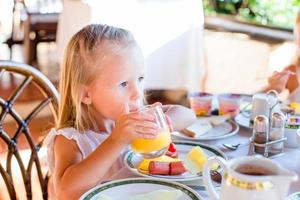 Adorable little girl having breakfast at restaurant. Cute kid enjoy fresh orange juice in outdoor cafe photo