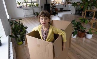 The girl hides in a big box and looks out of it funny in a room inside the home. Moving to a new house, unpacking things, renting an apartment, mortgage, housing issue photo