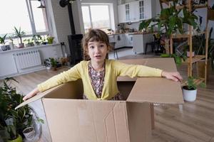 The girl hides in a big box and looks out of it funny in a room inside the home. Moving to a new house, unpacking things, renting an apartment, mortgage, housing issue