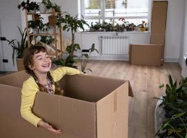 la niña se esconde en una caja grande y mira fuera de ella divertida en una habitación dentro de la casa. mudarse a una casa nueva, desempacar cosas, alquilar un apartamento, hipoteca, problema de vivienda foto