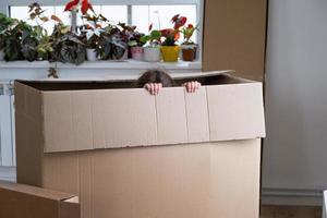 The girl hides in a big box and looks out of it funny in a room inside the home. Moving to a new house, unpacking things, renting an apartment, mortgage, housing issue photo